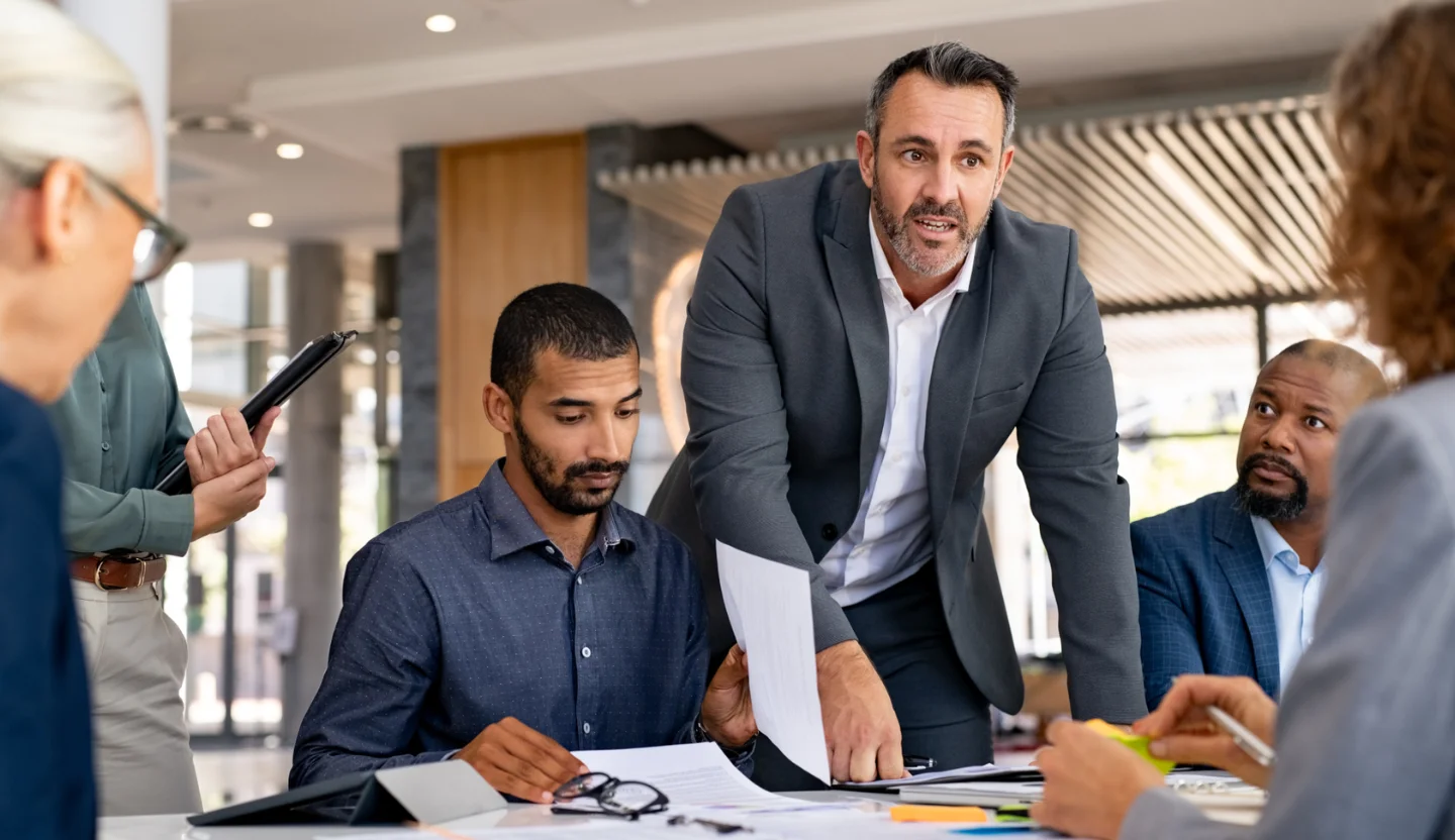 One office worker standing and leaning over a conference table while coworkers look on and listen