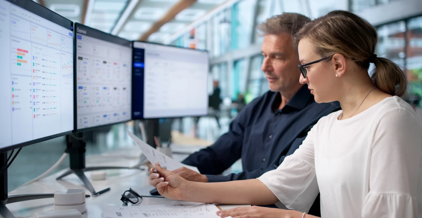Two office workers look at a document while at a table with multiple desktops
