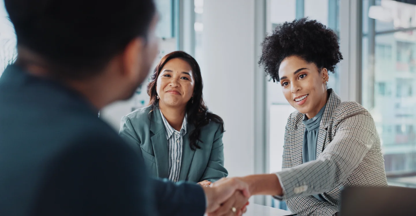 Two office workers shaking hands while another person looks on and smiles