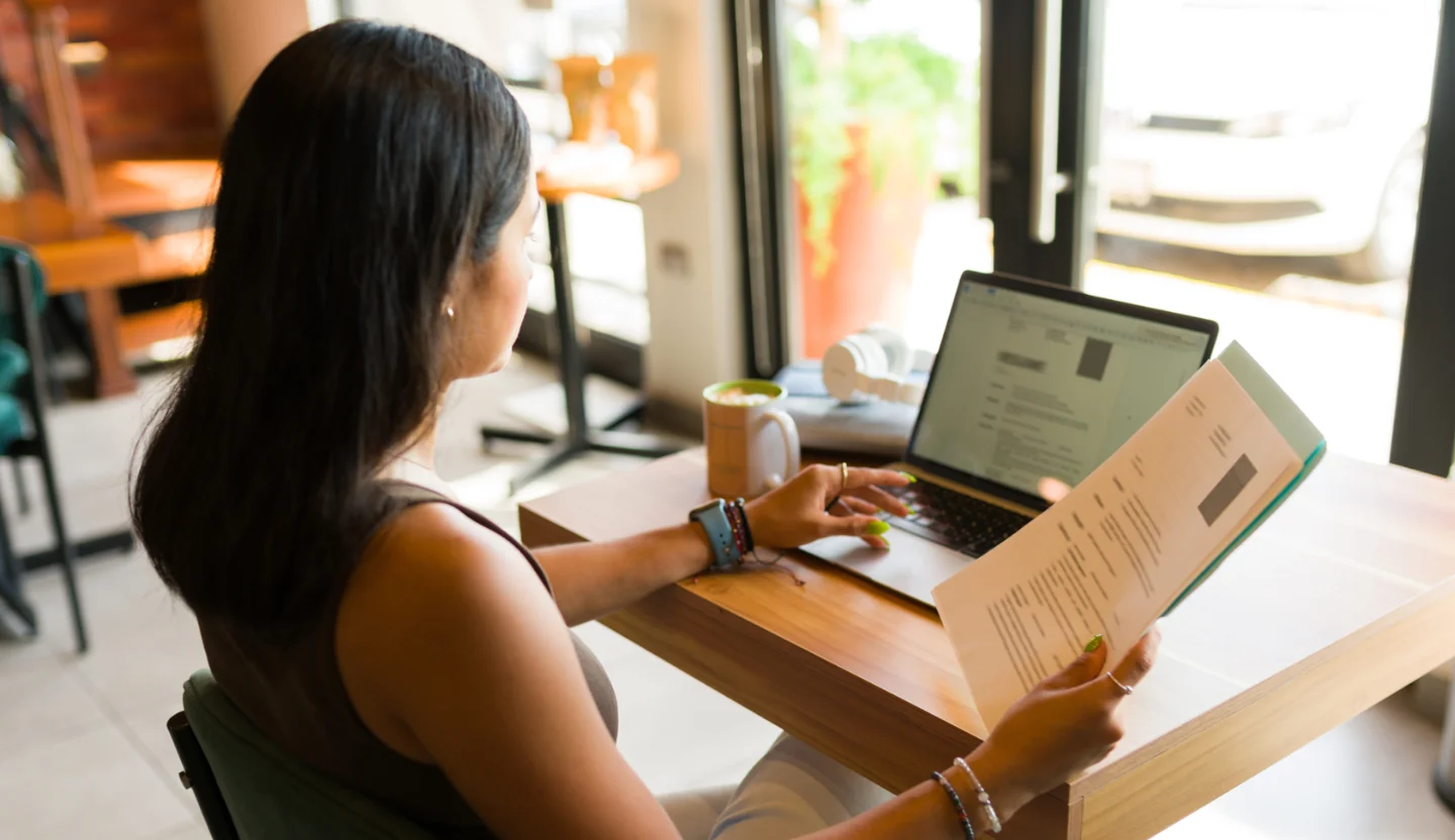 A person sits in front of a laptop while holding a piece of paper