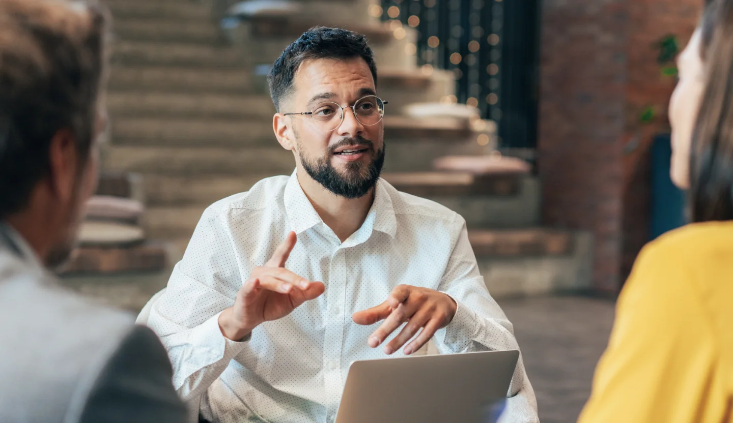 Business professional gestures while speaking with two colleagues around a laptop in a modern office